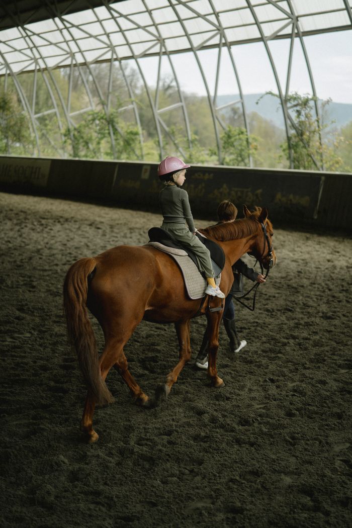 about-us-02 A young girl wearing a pink helmet is horseback riding in an indoor arena guided by an instructor.