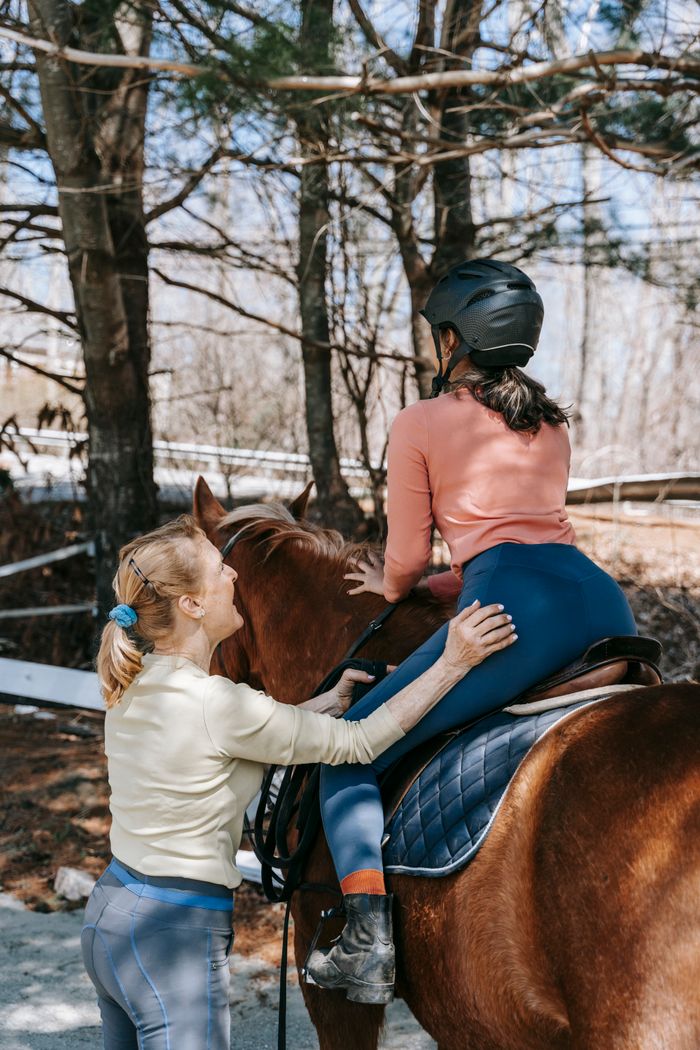 A woman helps a young girl on a horse, wearing equestrian gear in a forest setting.