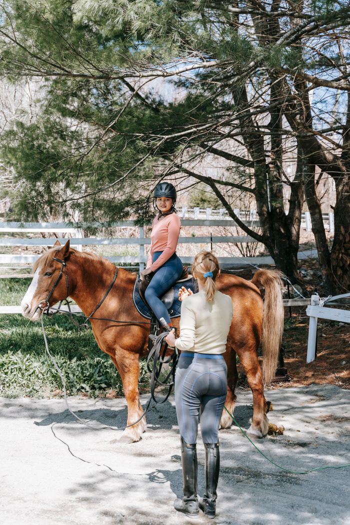 about-us-02 Two women involved in horseback riding training session outdoors in a wooded area.